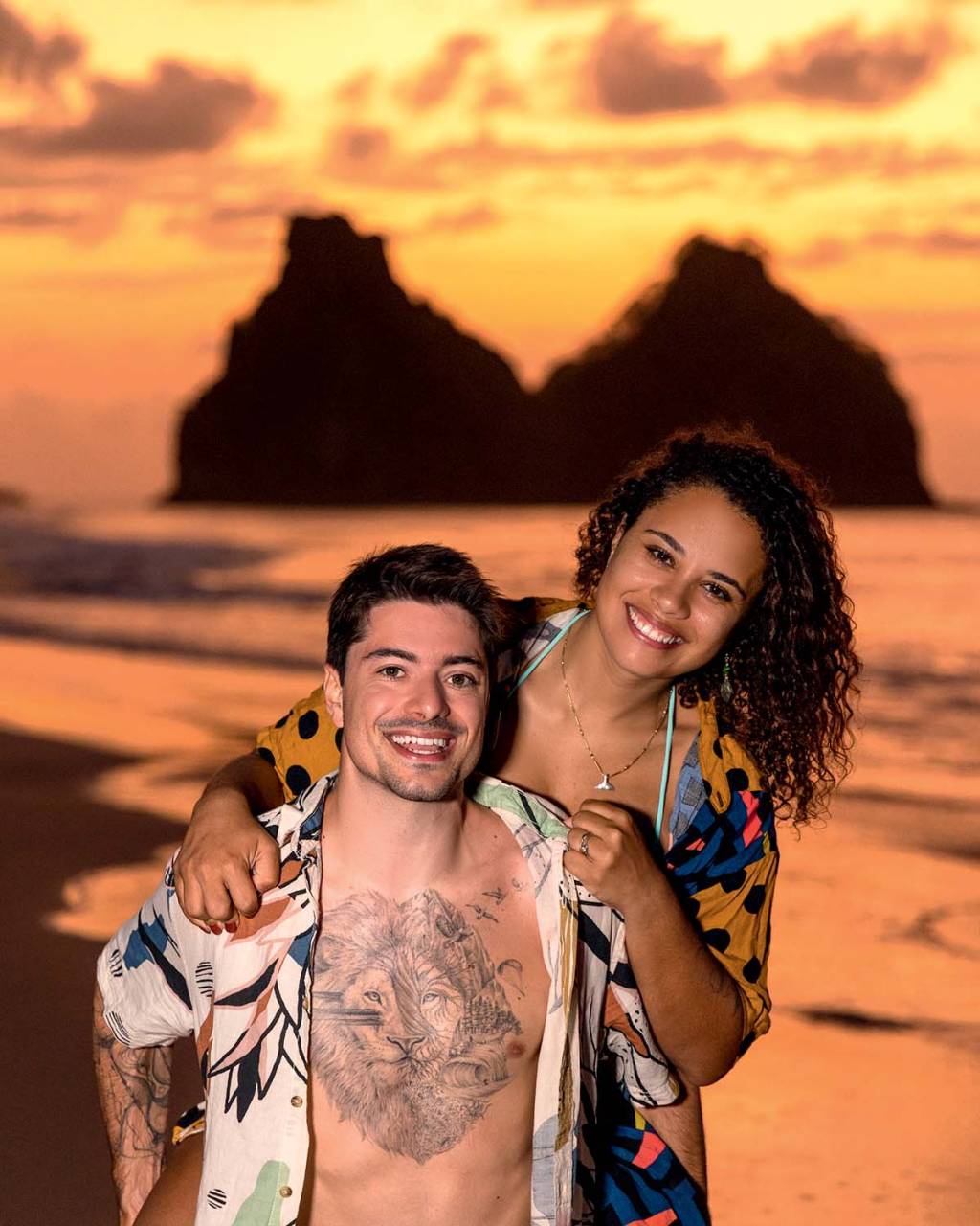 Homem branco de peito tatuado e camisa estampada posa com a esposa segurando seus ombros, uma mulher negra de cabelos cacheados. Posam com uma praia em tons alaranjados ao fundo.