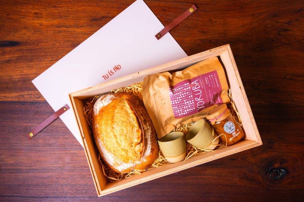 Foto aérea de caixa de madeira aberta sobre mesa de madeira com pão de fermentação natural, duas xícaras de cerâmica, pacote de café e vidro de geleia dentro.