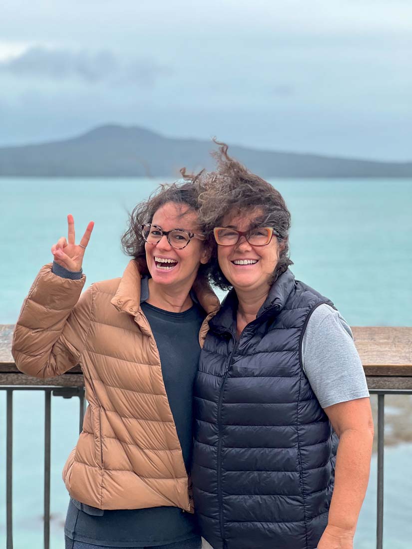 Duas mulheres brancas de óculos e cabelos esvoaçantes posam sorrindo em frente a mar verde.