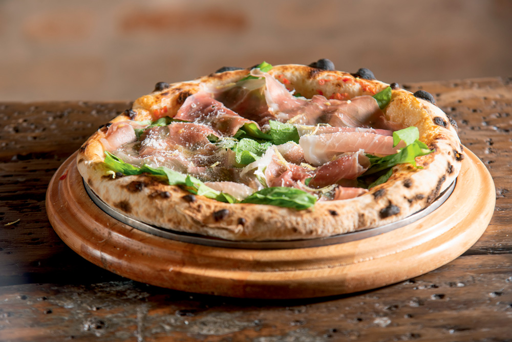 Pizza individual de bordas altas com presunto cru, parmesão, rúcula e raspas de limão-siciliano sobre madeira.