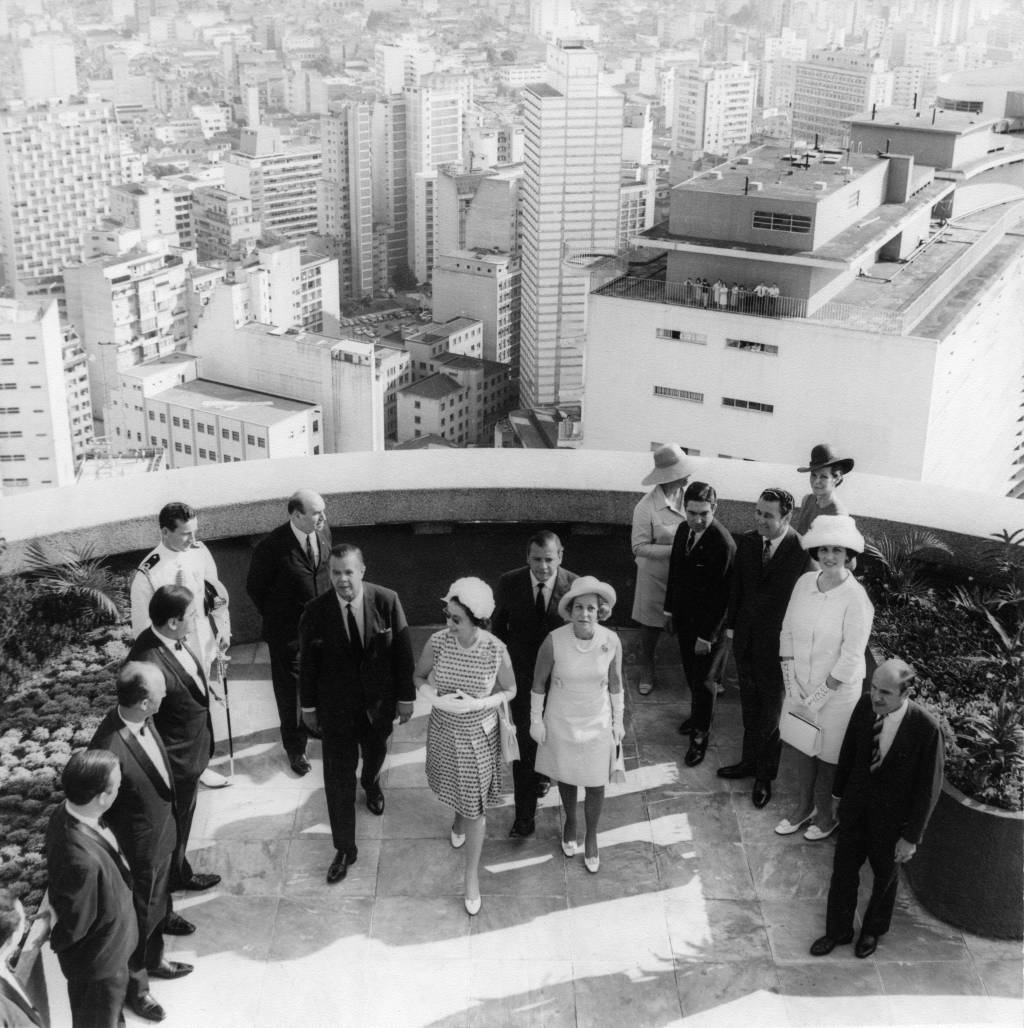 Foto em preto e branco exibe terraço de edifício com presença de convidados e rainha Elizabeth II no centro.