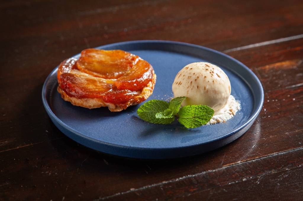 Prato azul sobre mesa de madeira com tarte tatin mini ao lado de bola de sorvete de creme enfeitada com ramo de hortelã.