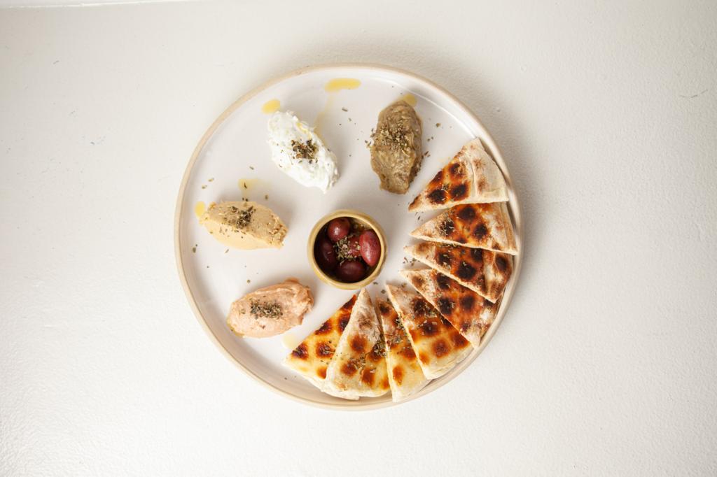 Foto aérea de mézzes, prato branco em cima de tampo branco com quatro pastas gregas de um lado, um potinho de azeitona ao centro e pães pita cortados em triângulo à direita.