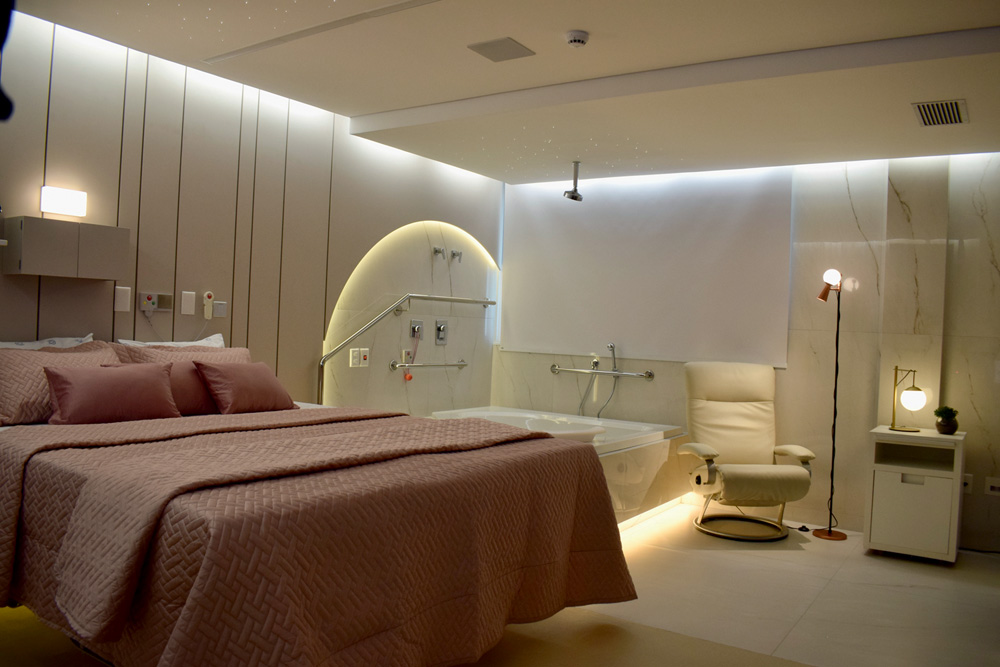 cama de casal Pro Matre – Divulgação.jpg Quarto de maternidade com cama de casal e iluminação nas bordas do local.