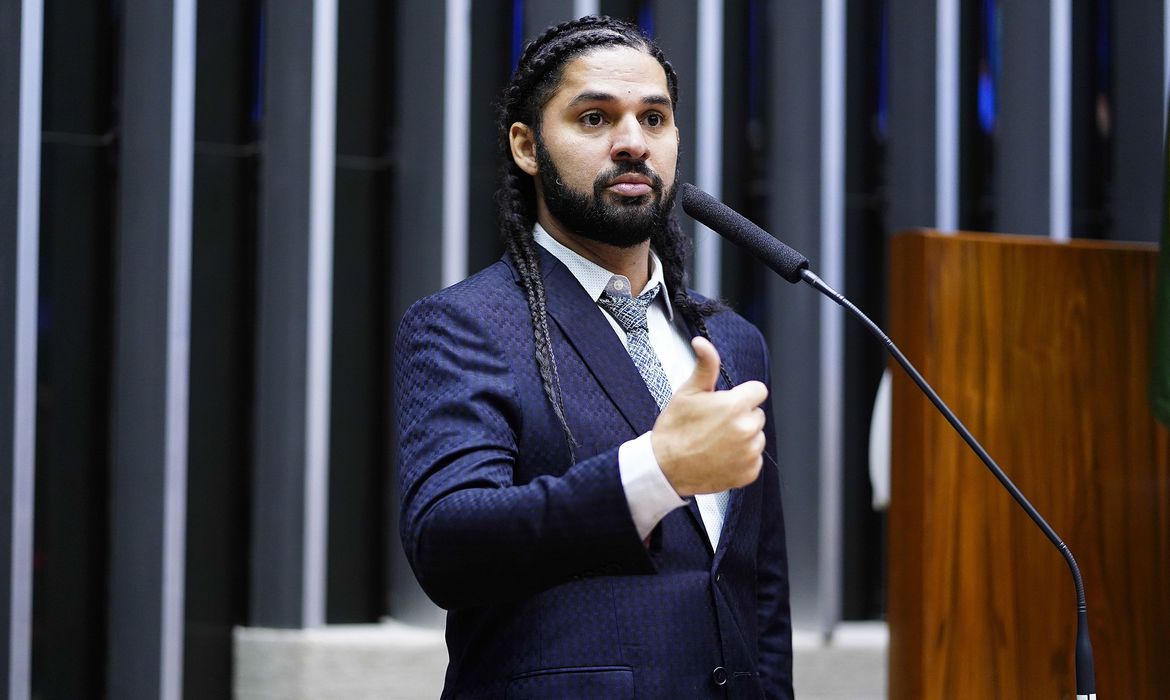 Um homem de terno, de tranças no cabelo, de pé, falando ao microfone