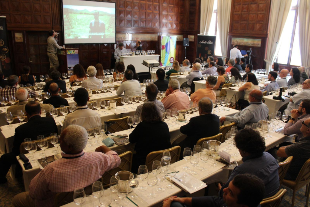 Rio Wine & Food Festival Pessoas de costas sentadas em mesas compridas alinhadas paralelamente cobertas por taças de vinho, assistindo apresentação em telão a frente.