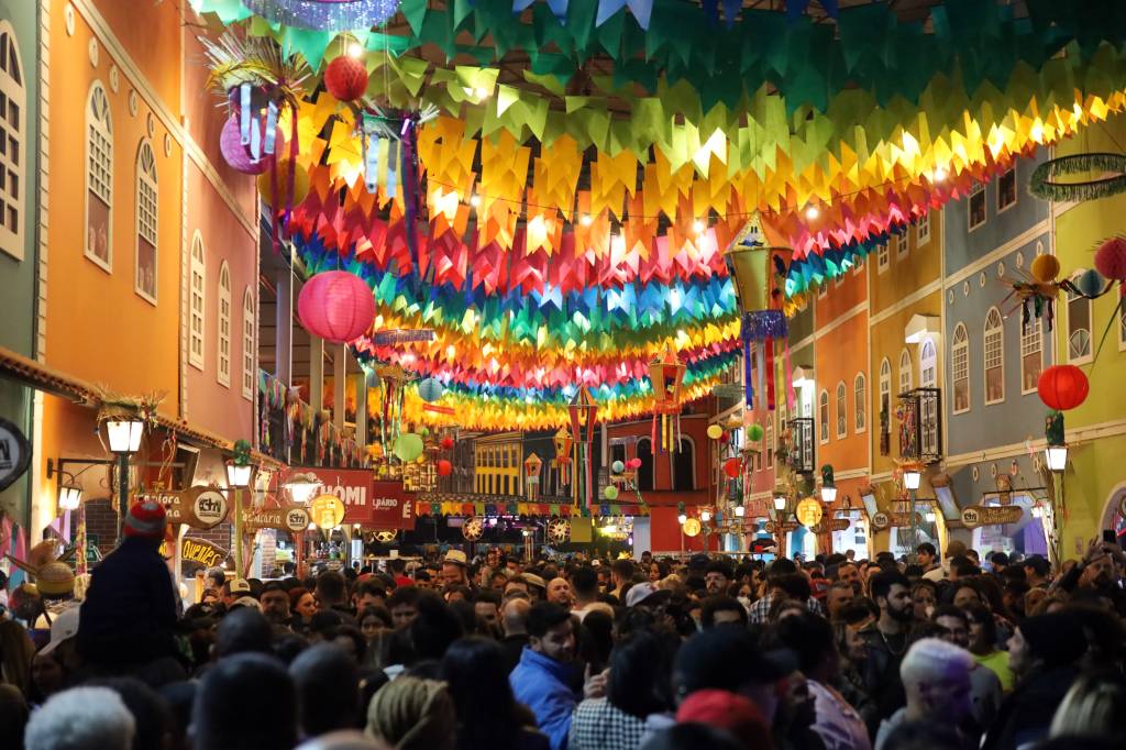 Grande corredor ocupado por muitas pessoas aparece adornado por fileiras de bandeirinhas coloridas e muitas luzes.