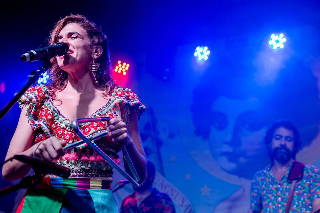 Cantora de cabelos castanhos lisos fala ao microfone de olhos fechados, em palco com luz e fundo azuis.