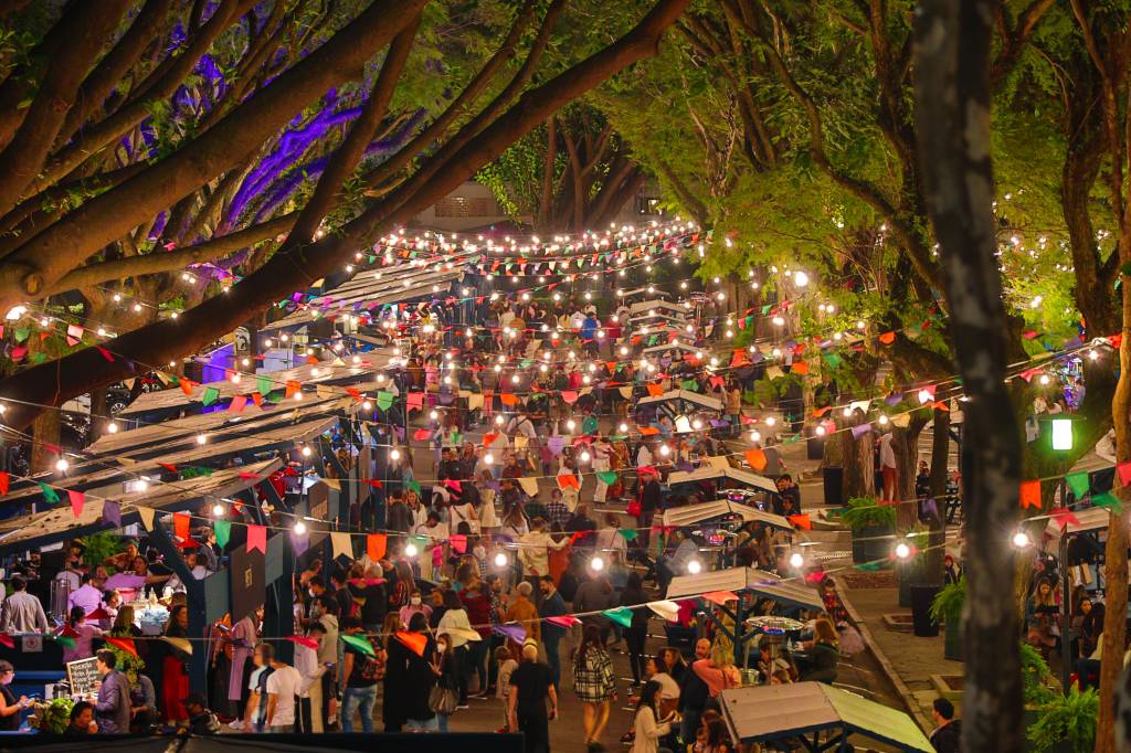 Foto exibe fileiras de bandeirinhas coloridas e iluminadas, abaixo pessoas circulando em rua fechada com pequenas tendas e árvores ao redor.