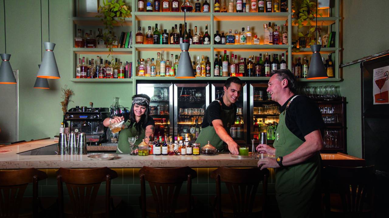 Uma mulher, à esquerda, e um homem, à direita, atrás de balcão de bar com um homem apoiado pelo lado de fora no lado direito.