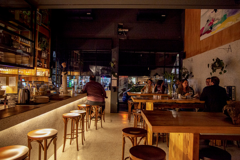 Salão em luz baixa do Elevado Bar com balcão iluminado com banquetas à esquerda e mesas altas à direita.