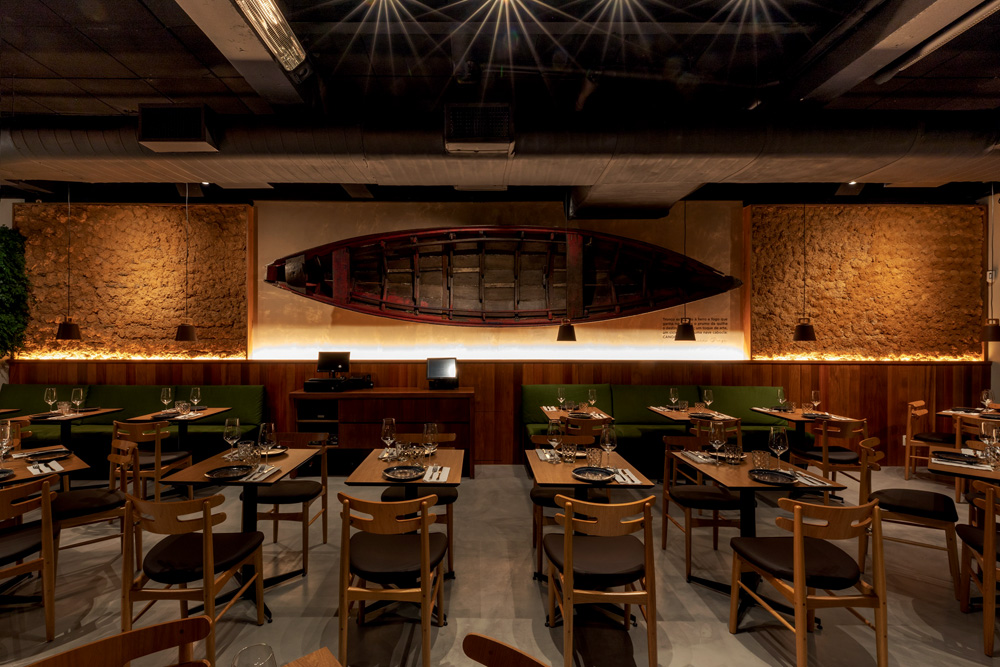Salão do restaurante Banzeiro com mesas e cadeiras de madeira e parede com canoa ao fundo.