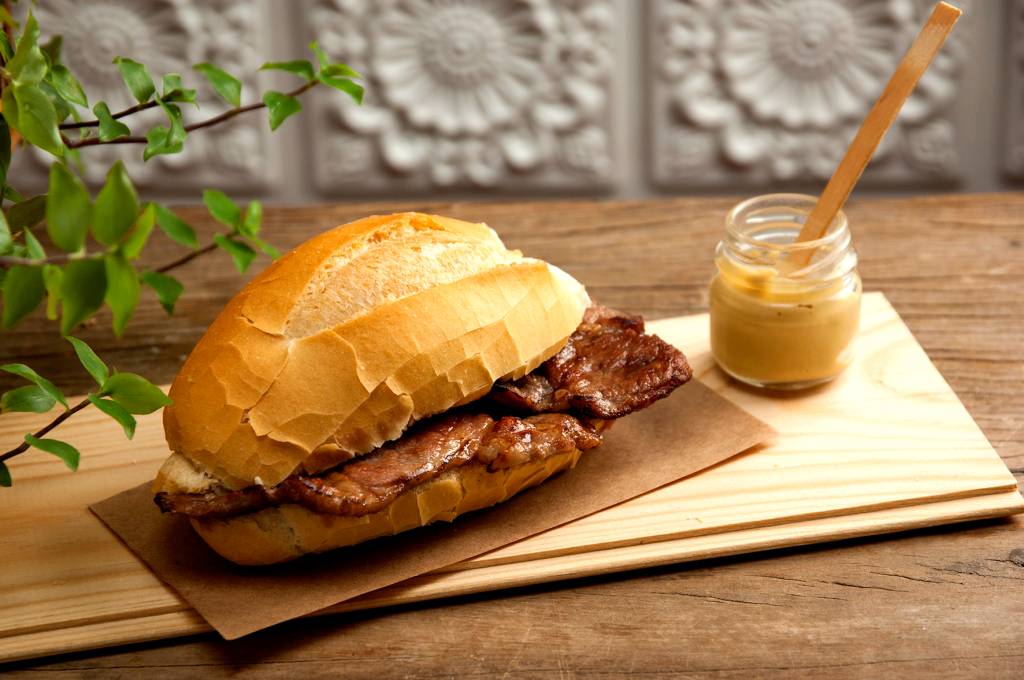 Sanduíche no pão francês sobre papel krafr com pedaço de carne aparecendo ao lado de potinho de maionese.