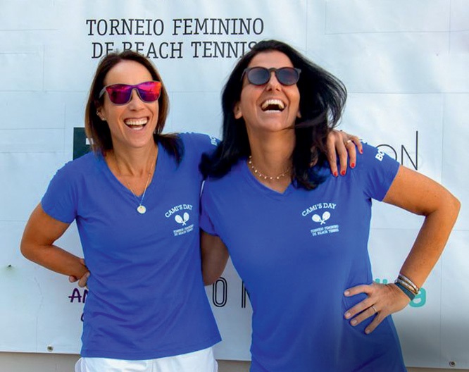 Duas mulheres brancas de cabelos pretos lisos sorriem abraçadas com camisetas azuis em frente a letreiro branco.