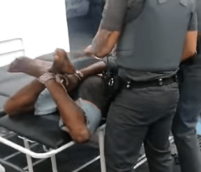 um homem negro com os pés e mãos atados por cordas sendo carregado por policiais