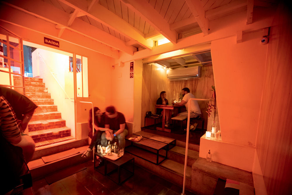 Salão do Cava Bar com luz alaranjada, escada à esquerda e casais sentados em mesas e bancos ao fundo.