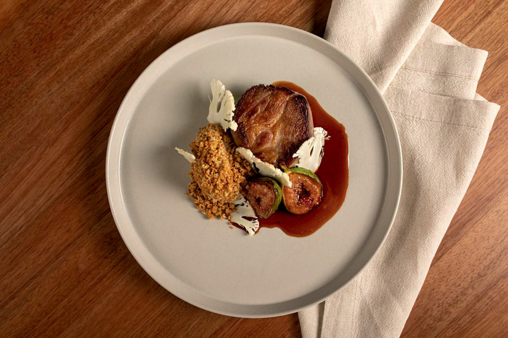 Foto aérea de prato de louça branca sobre mesa de madeira ao lado de guardanapo de pano branco com pedaço de porco, molho e figos.