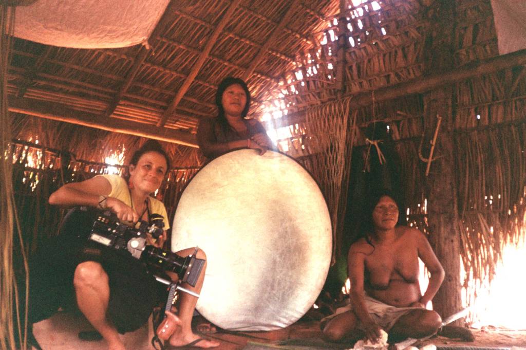 Mulher branca posa ao lado de duas mulheres indígenas dentro de cabana. Uma delas está de pé segurando um projetor de luz branco e redondo.