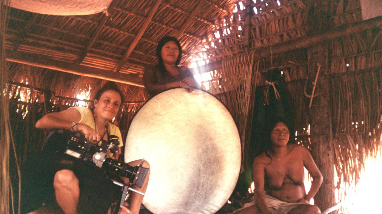 Mulher branca posa ao lado de duas mulheres indígenas dentro de cabana. Uma delas está de pé segurando um projetor de luz branco e redondo.
