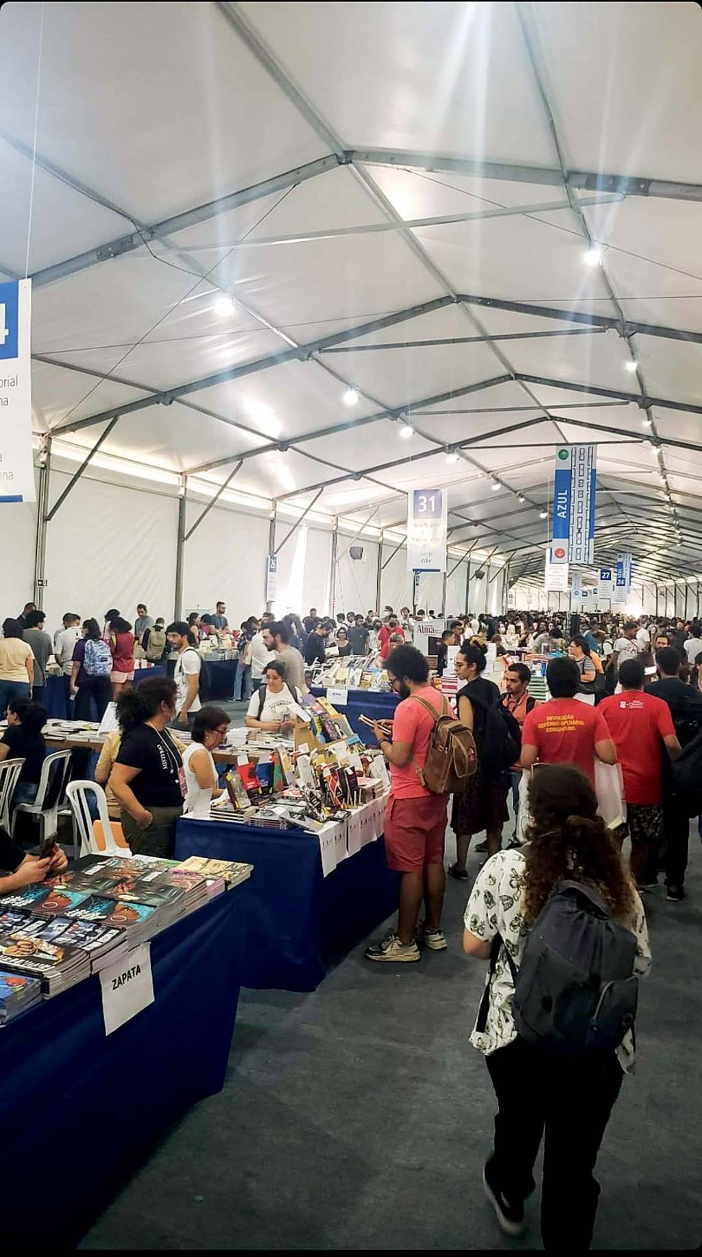 Foto exibe área interna de grande tenda com fileiras de mesas com livros e pessoas ao redor.