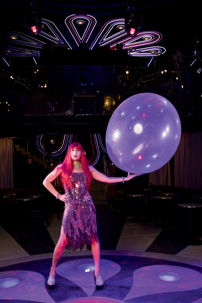 Uma pessoa com vestido prateado, máscara de rosto feminino humano e peruca vermelha posa com uma mão na cintura e a outra estendida segurando um balão transparente gigante.