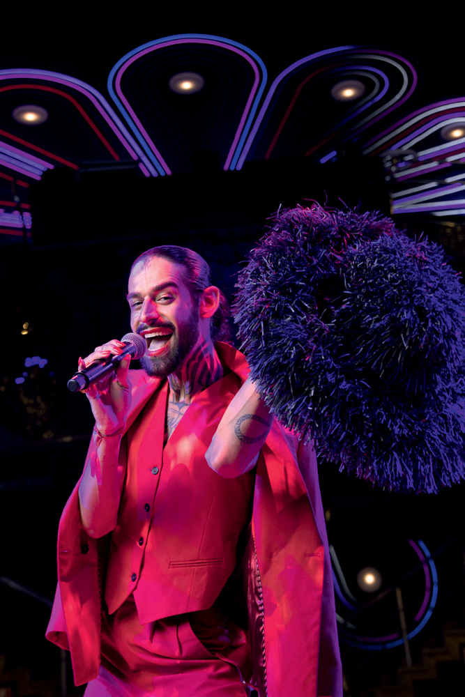 Travesti com cabelo preto preso e barba posa cantando em microfone e segurando um grande chapéu preto com material parecido com penas.