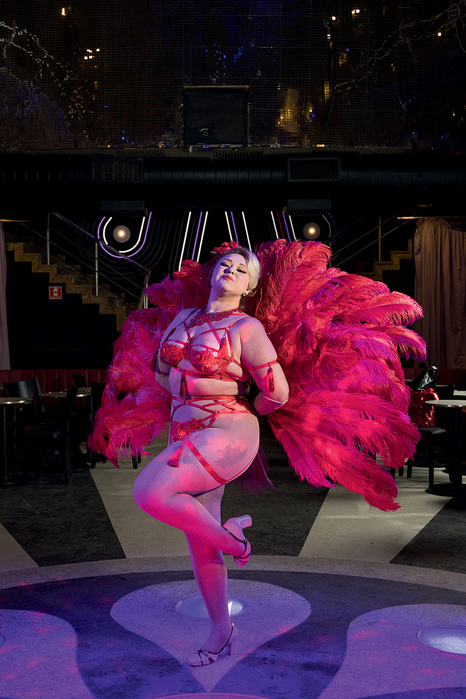 Mulher loira posa com uma das pernas dobrada, no centro de um palco circular preto e branco, segurando dois grandes leques vermelhos feitos de pluma atrás dela. Veste uma lingerie vermelha.