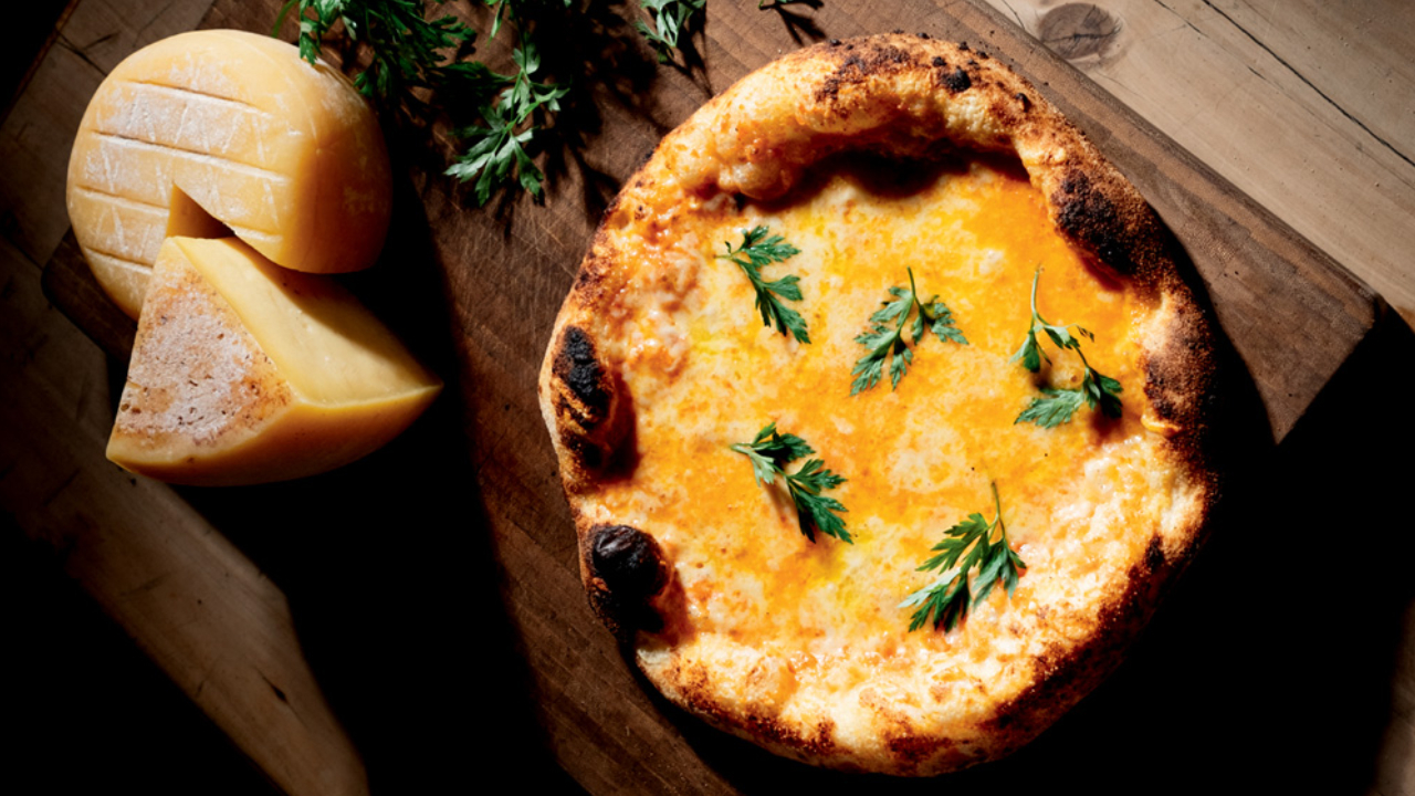 Pizza de queijo com folhas verdes sobre tábua de madeira ao lado de queijo maturado.