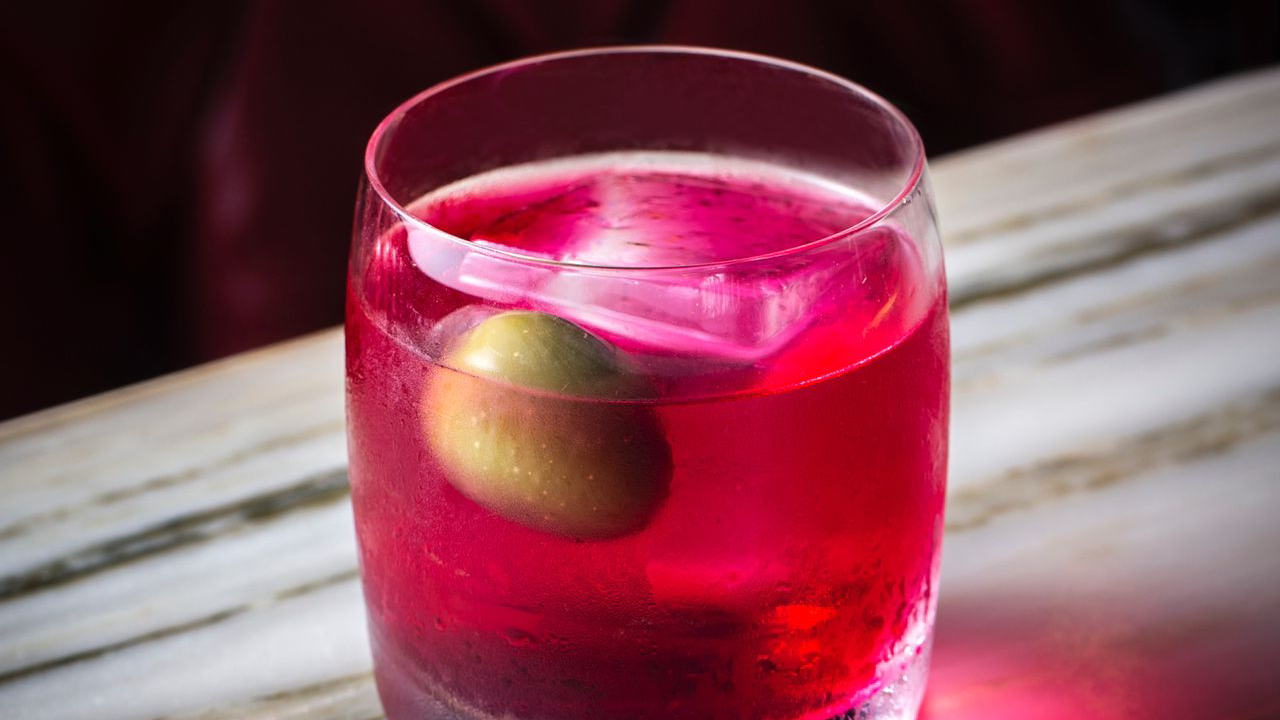 Drinque de copo baixo de cor rosa com azeitona verde dentro.