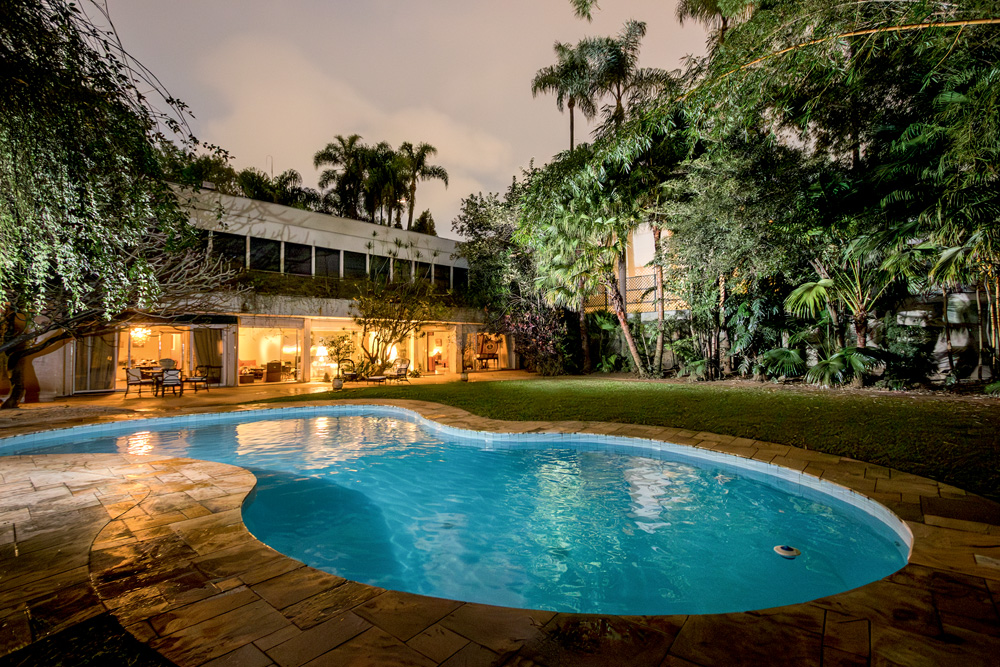 Piscina com borda oval aparece em primeiro plano ao lado de gramado e casa de dois andares em linhas retas.