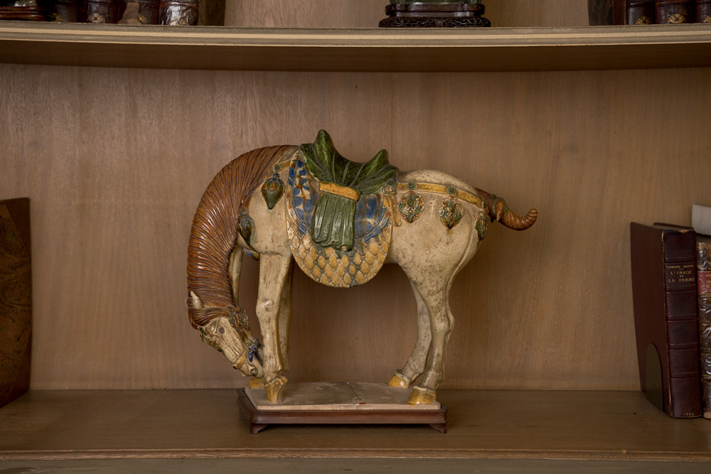 Cavalo de porcelana aparece de lado, com a cabeça abaixada, em prateleira de madeira.