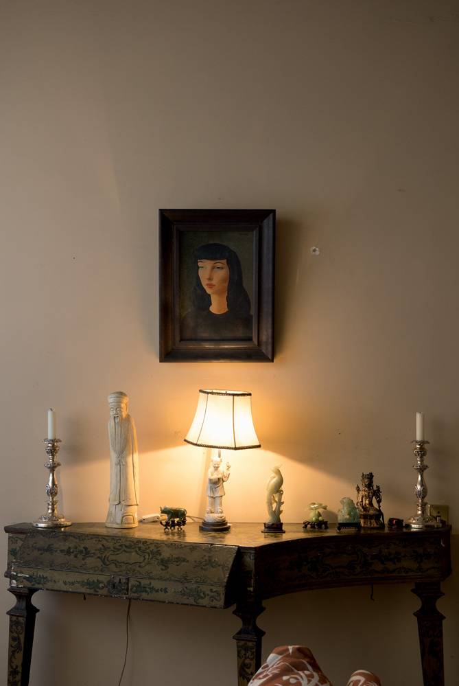 Parede com quadro de retrato de uma moça de cabelos pretos e mesa de canto com peças de marfim.