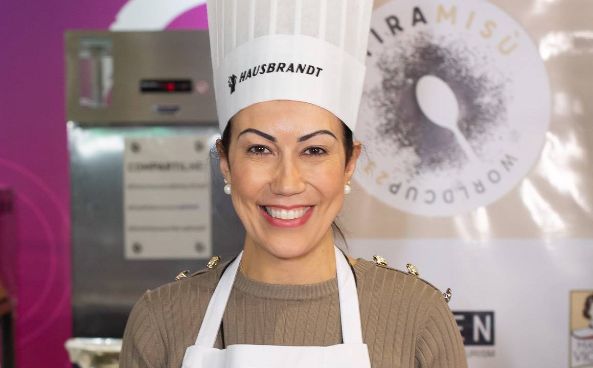 Mulher de avental branco e chapéu de cozinheira posa segurando prato com pavê italiano.