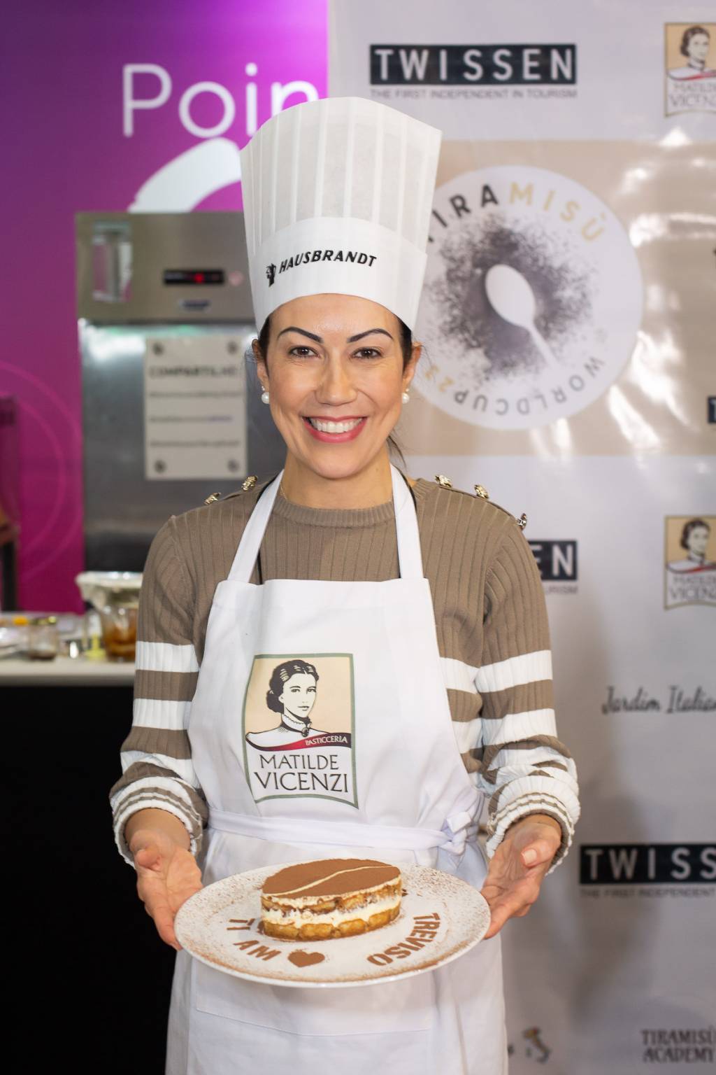 Mulher de avental branco e chapéu de cozinheira posa segurando prato com pavê italiano.