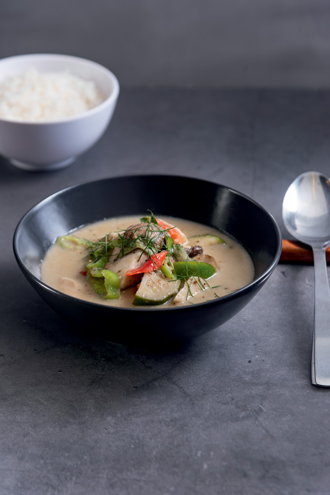 Cumbuca de louça preta com curry verde com legumes ao lado de pote de arroz e colher.