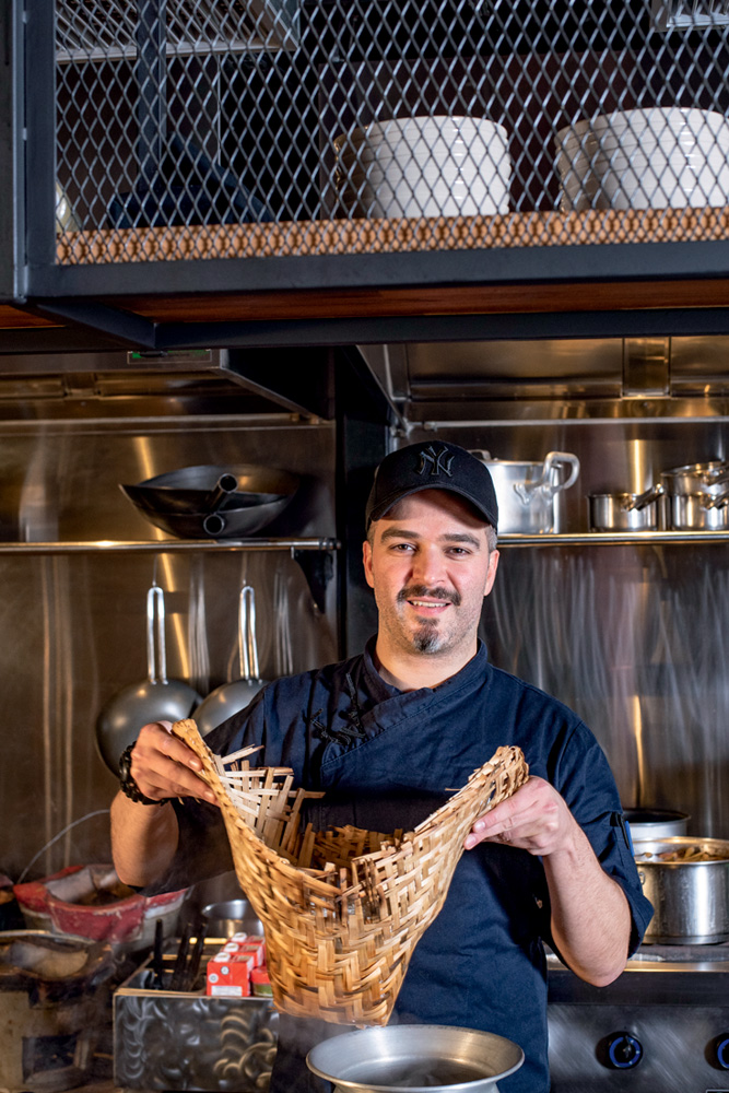 O chef Maurício Santi de boné e avental segurando cesta de palha na cozinha do Ping Yang.