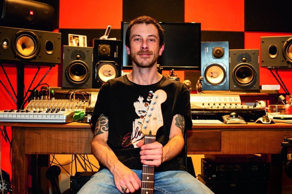 Homem branco de bigode posa segurando guitarra em estúdio. Veste calça jeans e camiseta preta.