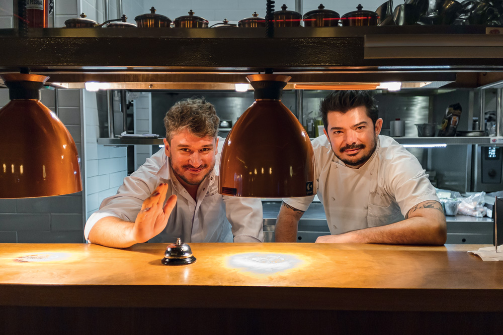 Dois chefs brancos de dólmã atrás de boqueta em cozinha de restaurante.