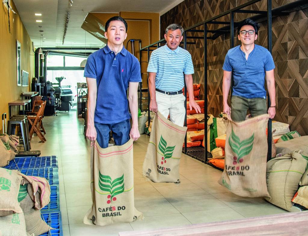 Três homens asiáticos fazendo corrida do saco com sacos de café dentro de uma cafeteria.