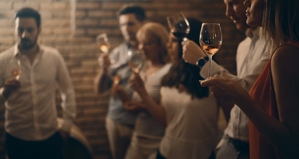 Pessoas alinhadas segurando taças de vinhos variados em prova.