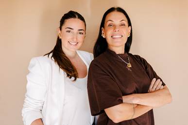 Duas mulheres brancas posam lado a lado, a da esquerda veste roupas brancas e a da direita veste camiseta de manga curta na cor marrom. Ela posa de braços cruzados. Ambas sorriem.