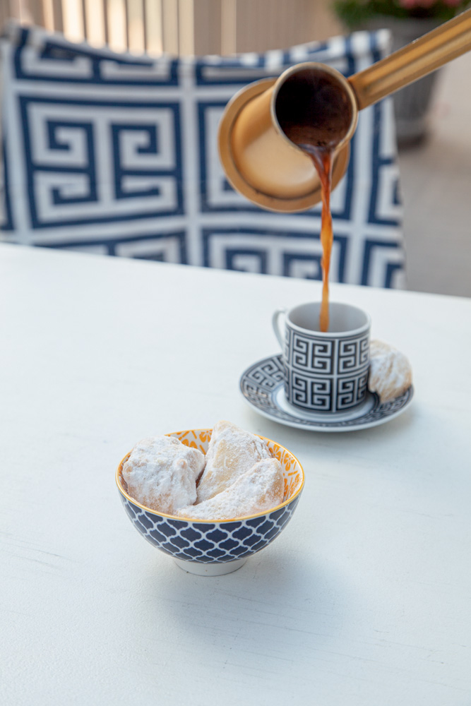 Café sendo servido de jarra de cobre ao fundo e cumbuca de louça azul com biscoitos envoltos em açúcar.