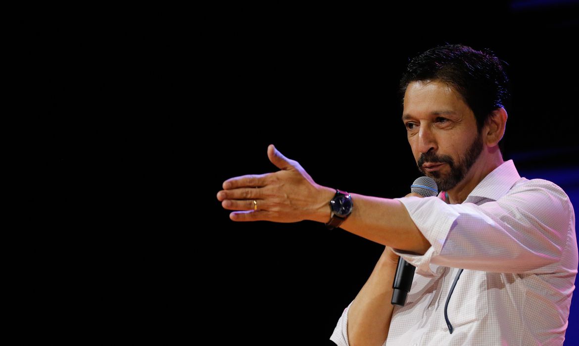Ricardo Nunes vestindo uma camisa social branca, com um fundo preto, enquanto fala com o público utilizando um microfone e aponta com o braço esquerdo na direção do público.