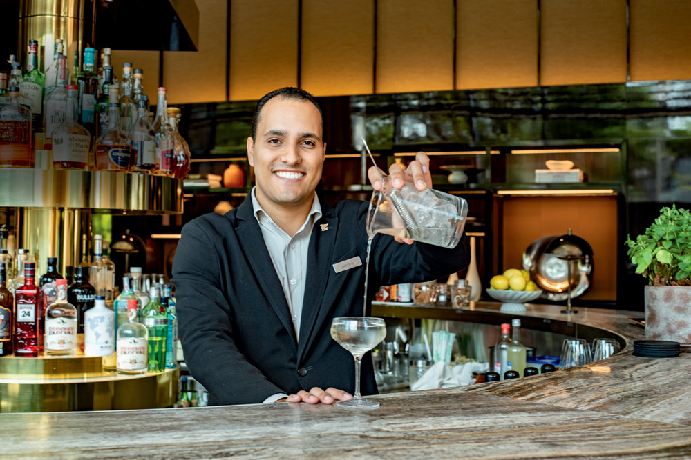 Gustavo Rômulo, bartender do Bar Caju, está sorrindo para a foto enquanto segura jarra de drinque que está sendo despejado na taça.