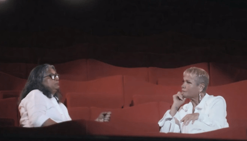 Marlene Mattos e Xuxa sentadas uma de frente pra outra em cadeiras de um teatro