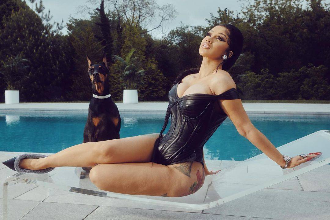 Mulher negra posa à beira de piscina com espartilho preto e cachorro de grande porte preto ao lado.
