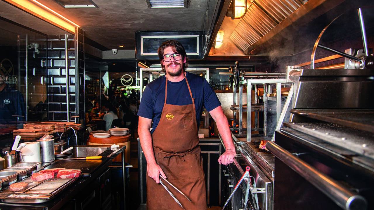 O churrasqueiro Renzo Garibaldi de avental segurando uma pinça dentro de cozinha industrial.
