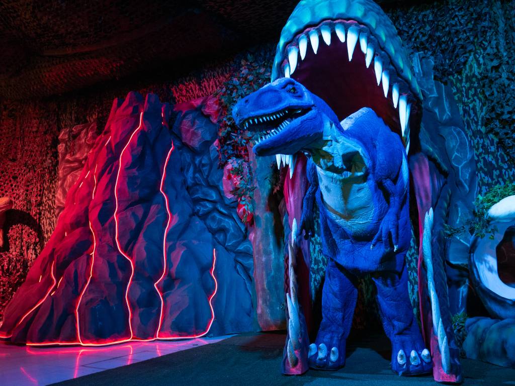 Ambiente do Magic Dining Dinos. Um boneco de dinossauro pousa dentro de um portal feito de uma grande cabeça de dinossauro inflável. À esquerda, há um vulcão cenográfico iluminado por leds laranja. O ambiente está escuro, o que faz com que tudo esteja meio azul escuro