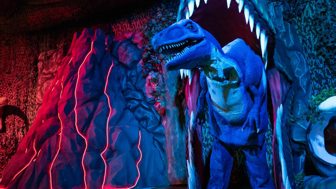 Ambiente do Magic Dining Dinos. Um boneco de dinossauro pousa dentro de um portal feito de uma grande cabeça de dinossauro inflável. À esquerda, há um vulcão cenográfico iluminado por leds laranja. O ambiente está escuro, o que faz com que tudo esteja meio azul escuro
