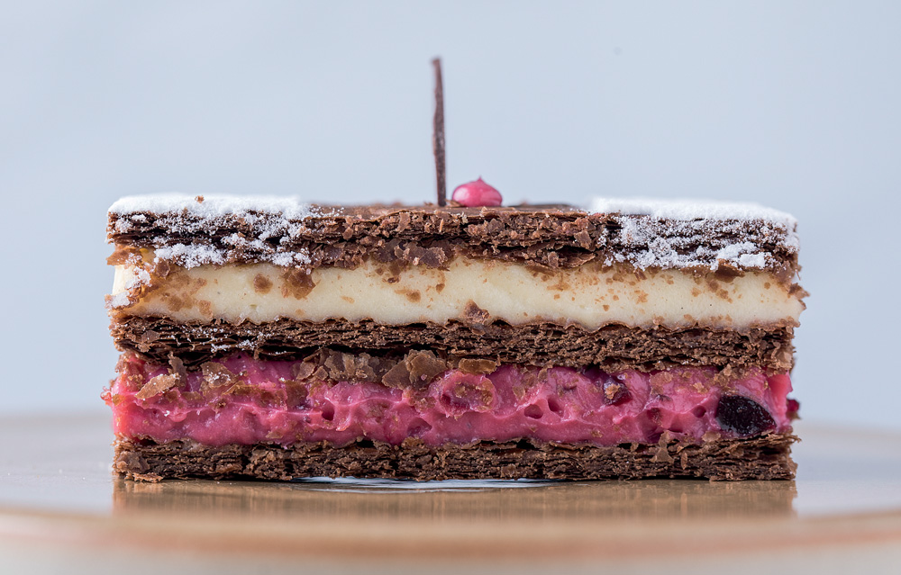Imagem frontal de mil folhas com massa de chocolate, uma camada de recheio branco e outra de recheio rosado.